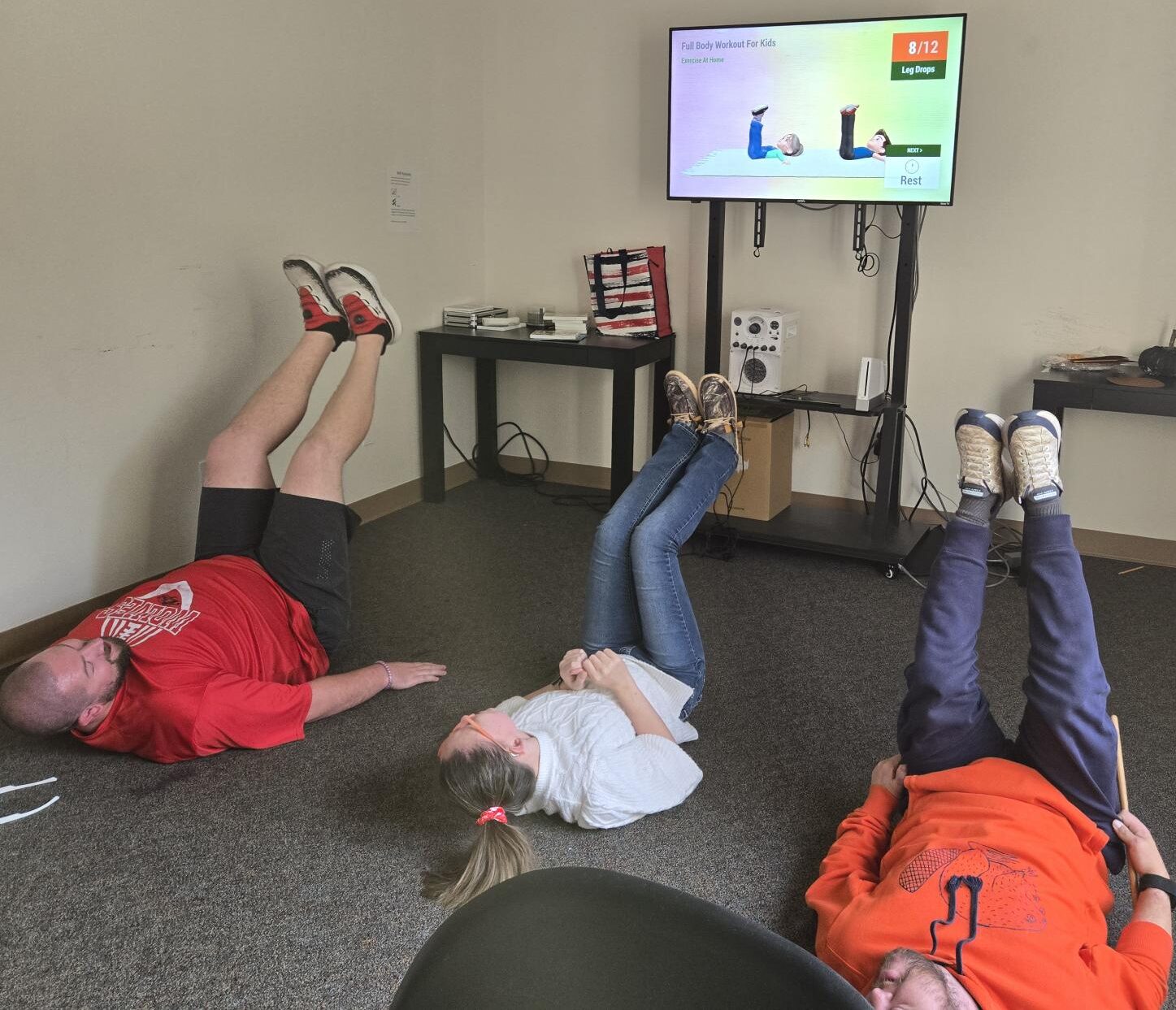 Three adults laying on the floor in a room, exercising to a video on the TV in front of them.