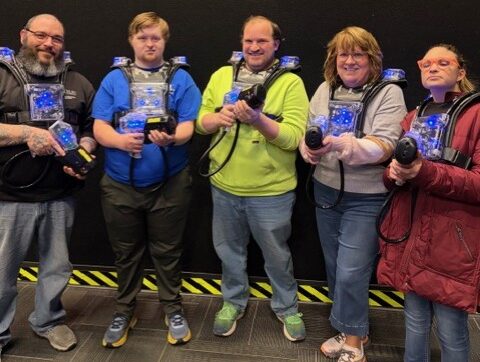 Group of five adults smiling, dressed in paintball attire holding paintball guns.
