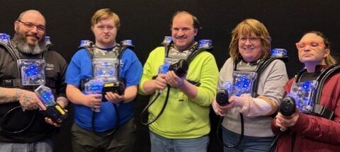 Group of five adults smiling, dressed in paintball attire holding paintball guns.